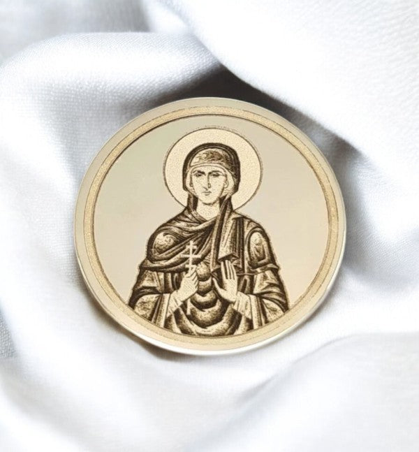 Zlatnik sa gravurom Svete Petke, prikazane sa oreolom, u mirnoj pozi, sa rukama na grudima koje drže krst i svitak, na glatkoj pozadini sa svetlom postavkom tkanine u pozadini.