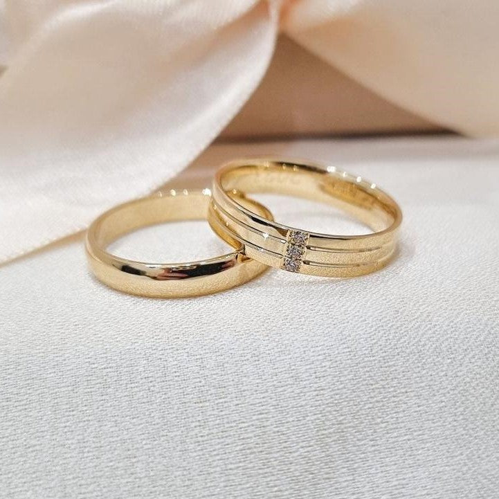 Zlatne burme od žutog zlata, jedna klasična i glatka, druga sa urezanim linijama i cirkonima u sredini, postavljene na bež tkanini sa svetlom pozadinom.