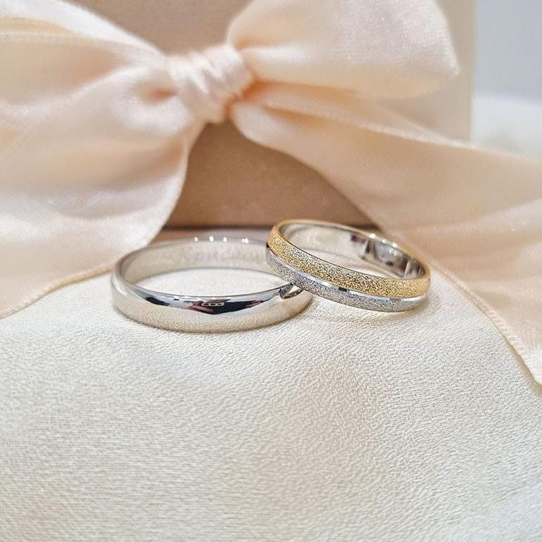 Par burmi od belog i žutog zlata – jedna klasično sjajna, druga sa mat zlatnim detaljem, položene na svetloj tkanini ispred satenske mašne u bež nijansi.