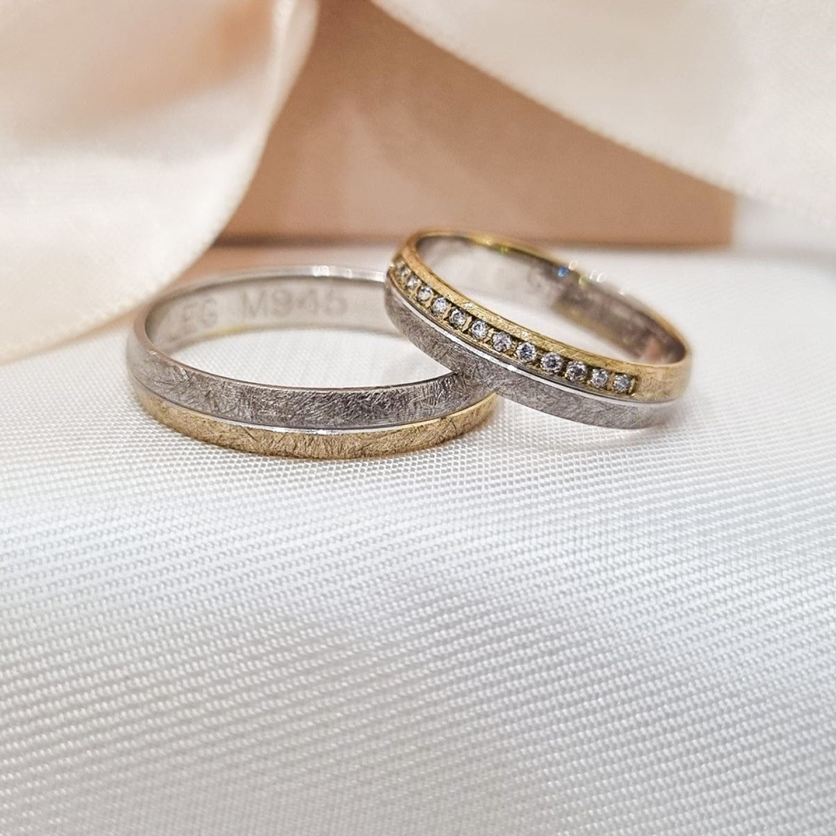 Par burmi od belog i žutog zlata – jedna sa mat finišem, a druga sa cirkonima duž gornje ivice, položene na svetloj tkanini ispred satenske mašne.