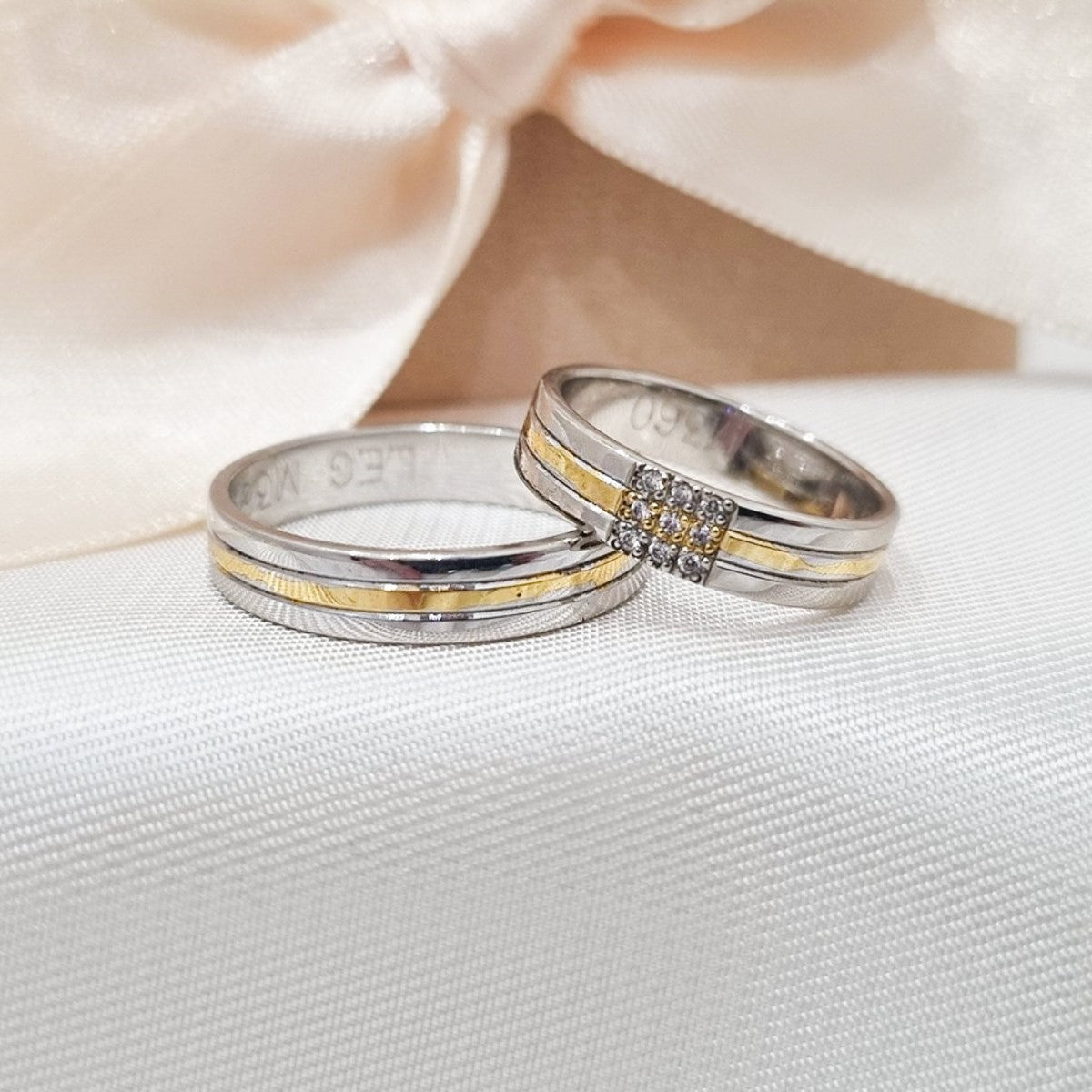 Elegantne burme od belog i žutog zlata, jedna ukrašena sitnim kamenčićia, položene na beloj svilenoj podlozi sa svetlom mašnom u pozadini.