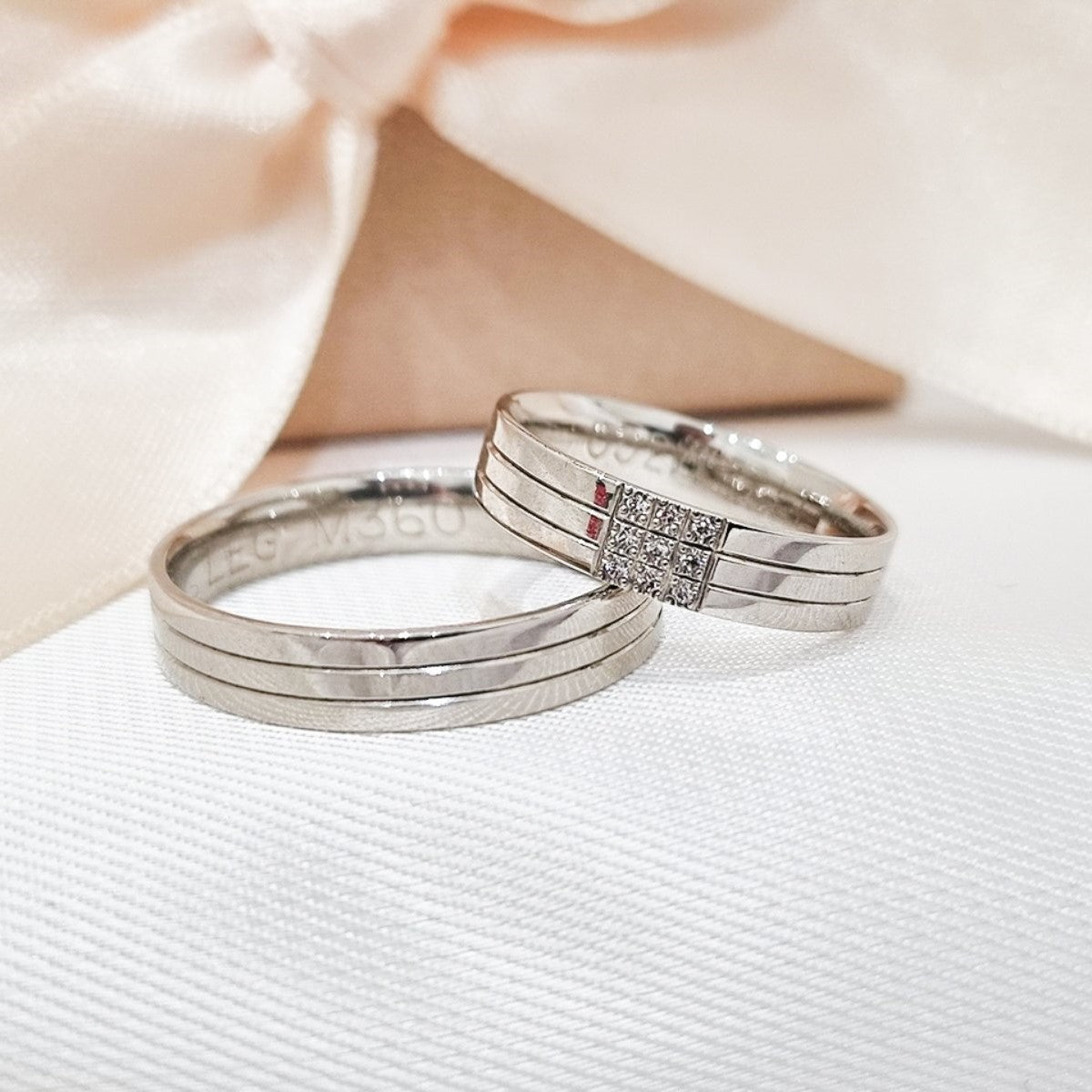 Zlatne burme sa tri fine linije i ukrasom od cirkona na jednoj, postavljene na bež tkanini sa svetlom pozadinom.