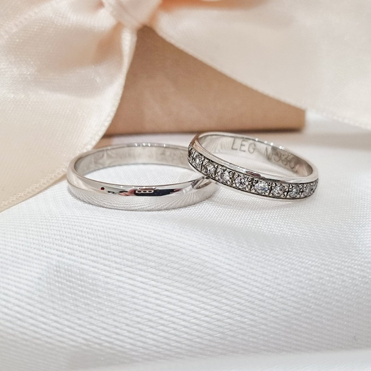 Set burmi od belog zlata – muška glatka burma i ženska burma sa redom ugrađenih cirkona, postavljene na beloj tkanini.