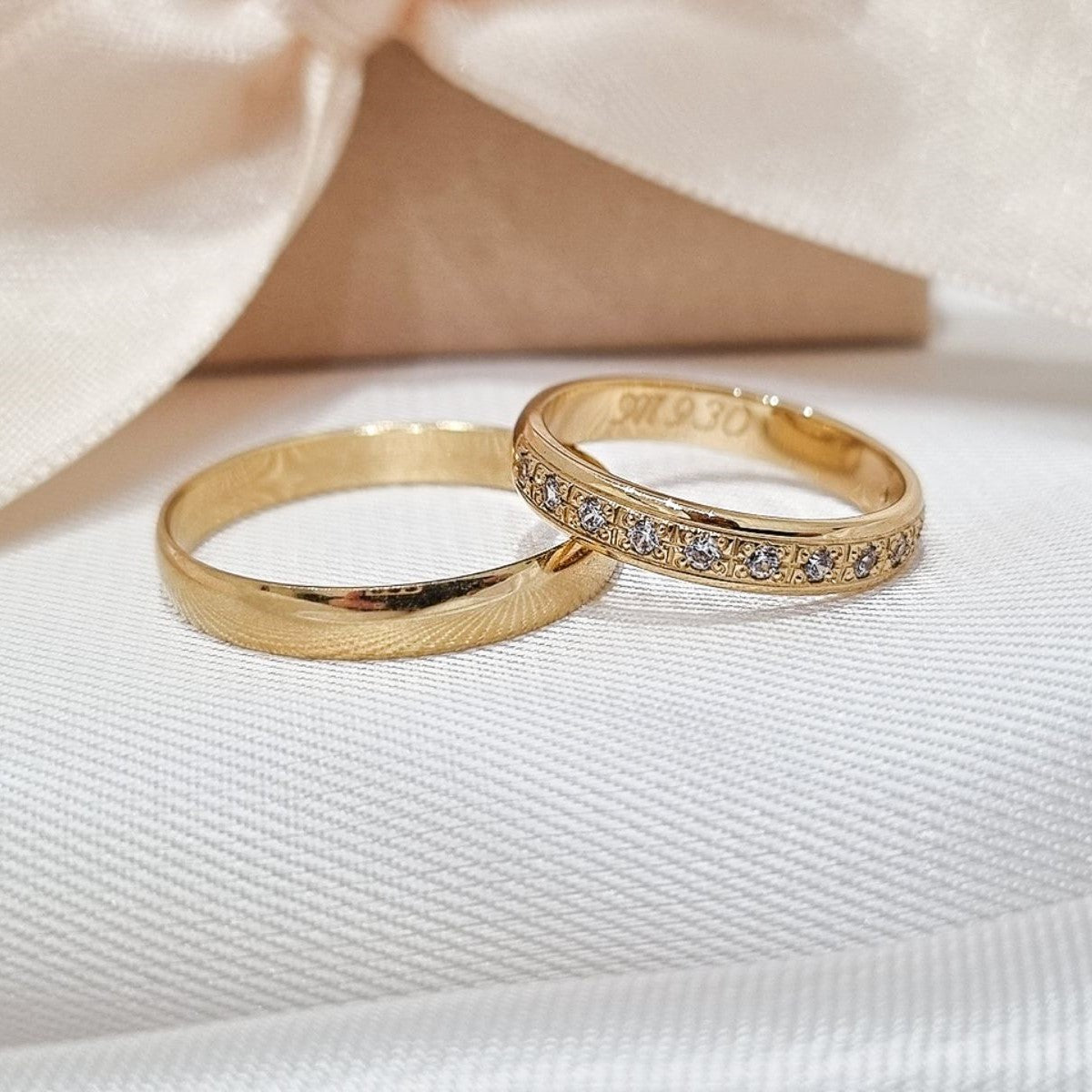 Zlatne burme, jedna sa poliranom i matiranom površinom, dok je druga ukrašena cirkonima, postavljene na bež tkanini sa svetlom pozadinom.