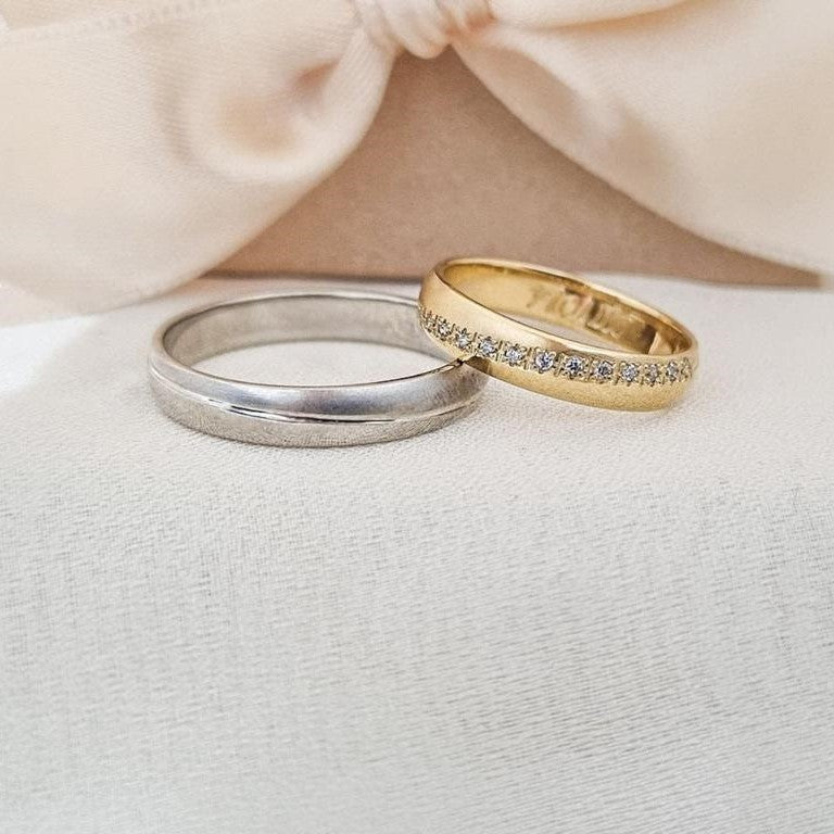 Burme od belog i žutog zlata – jedna sa mat efektom, druga sa cirkonima u nizu, položene na bež tkanini ispred satenske mašne.