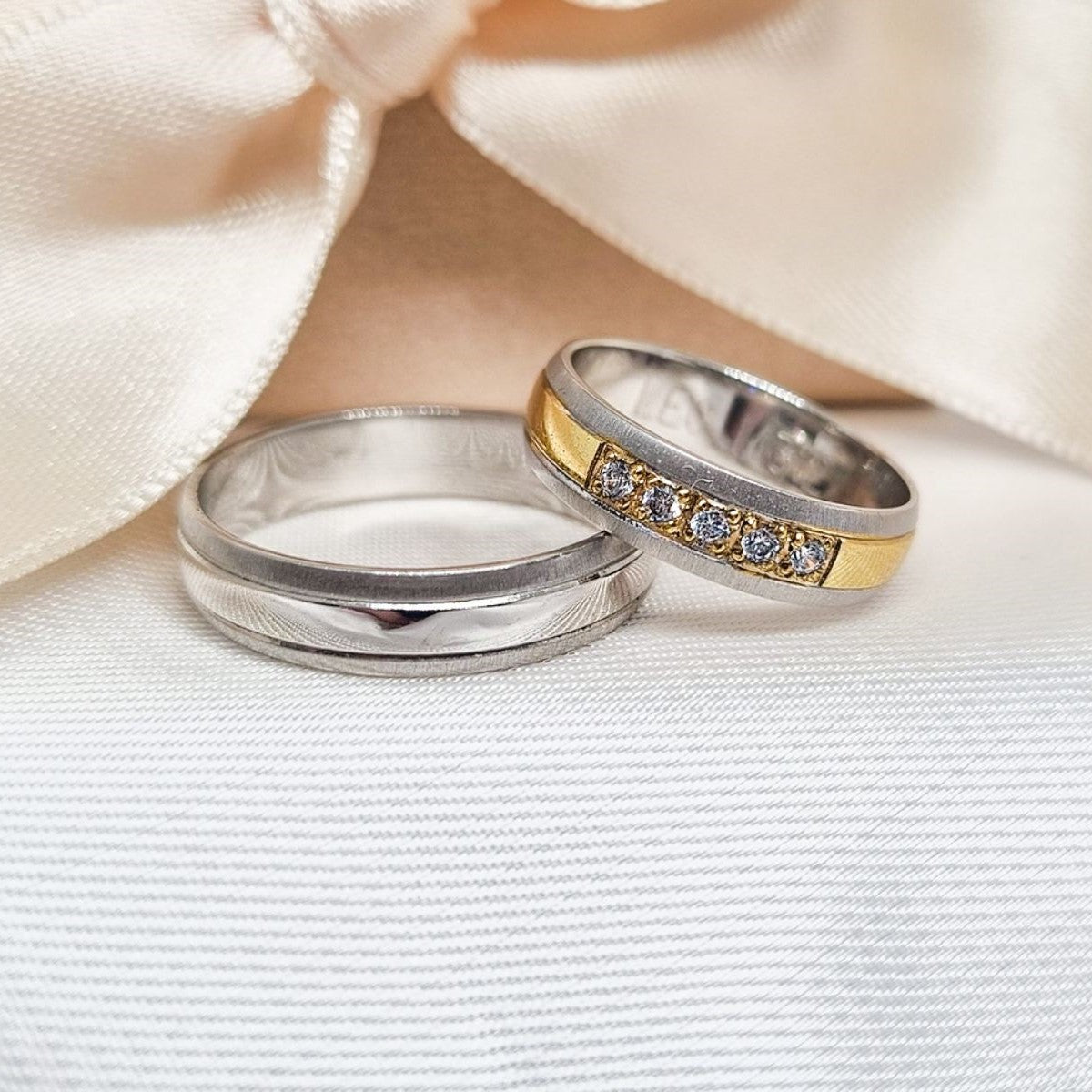 Zlatne burme sa kombinacijom belog i žutog zlata, jedna polirana, a druga ukrašena cirkonima, postavljene na bež tkanini sa svetlom pozadinom.