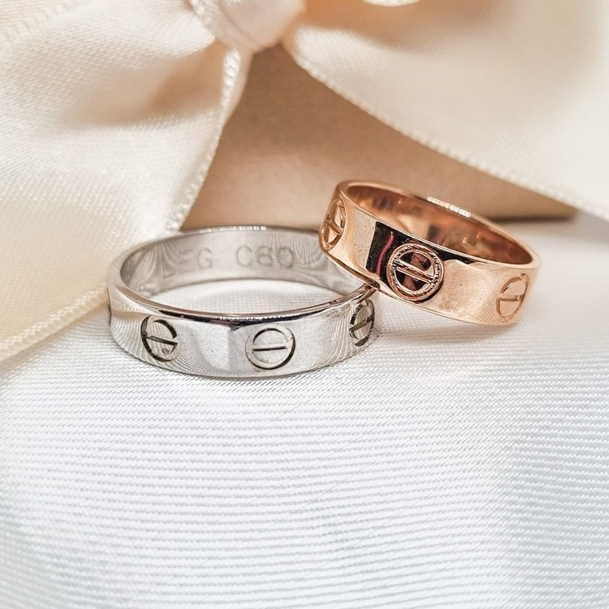 Zlatne burme u kombinaciji ružičastog i belog zlata sa ugraviranim simbolima, postavljene na bež tkanini sa svetlom pozadinom.