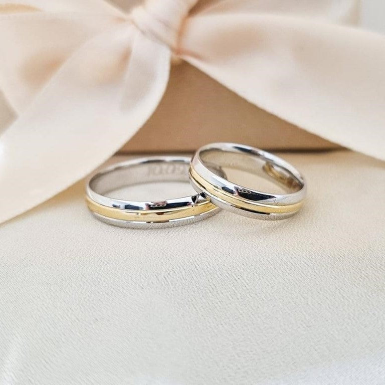 Dve burme od belog i žutog zlata sa sjajnom završnom obradom, položene na bež površini ispred satenske mašne.