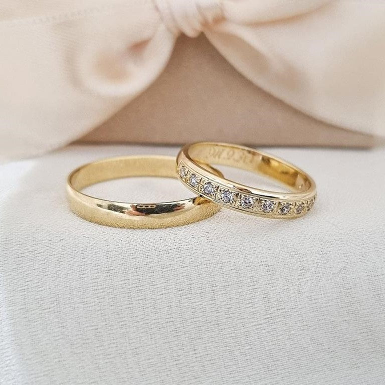 Burme od žutog zlata – jedna sa matiranom završnicom, druga sa nizom svetlucavih cirkona, postavljene na bež podlozi ispred svilenkaste mašne.