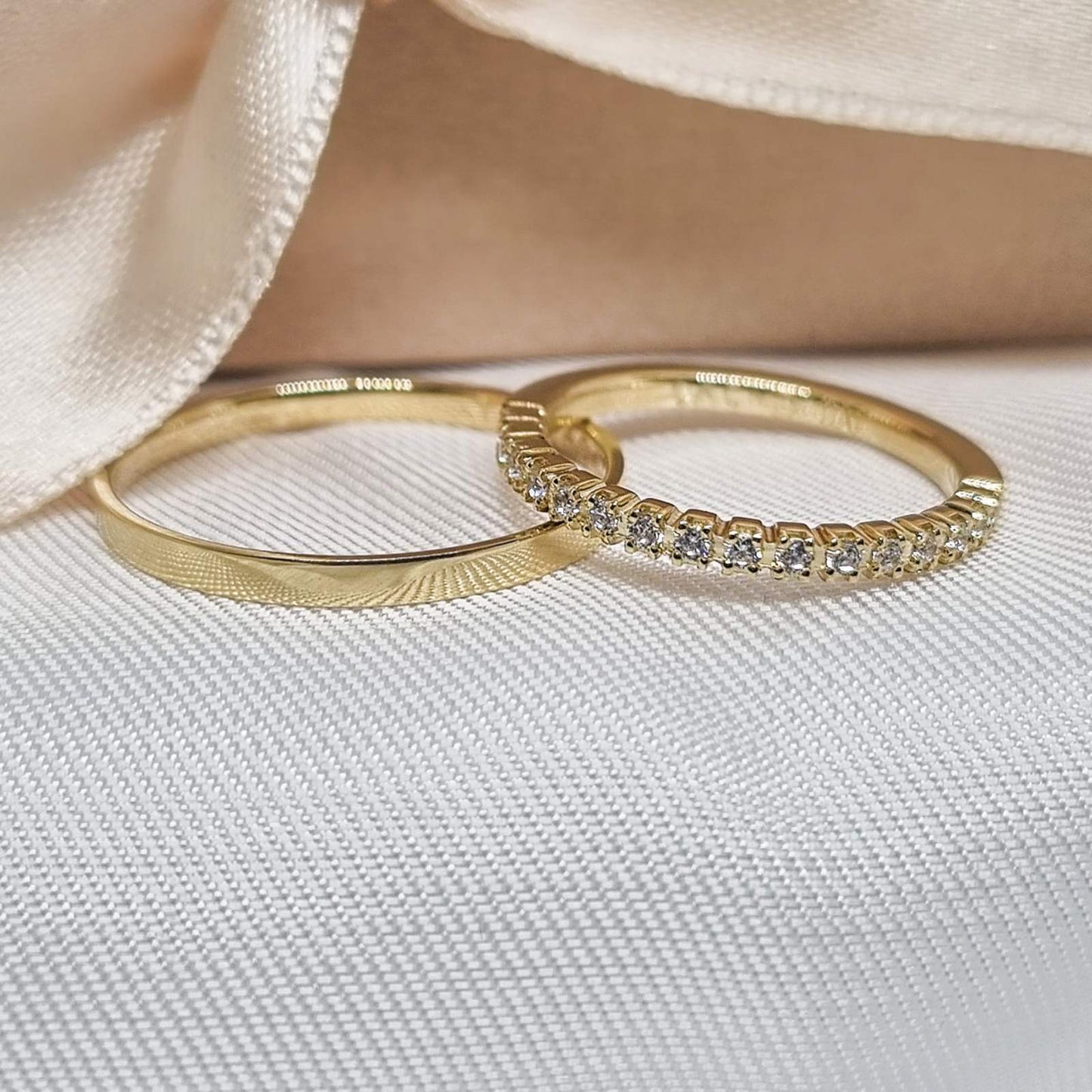 Par burmi od žutog zlata – jedna ukrašena redom sitnih cirkona, druga glatka s diskretnom teksturom, na beloj tkanini uz svilenu mašnu.