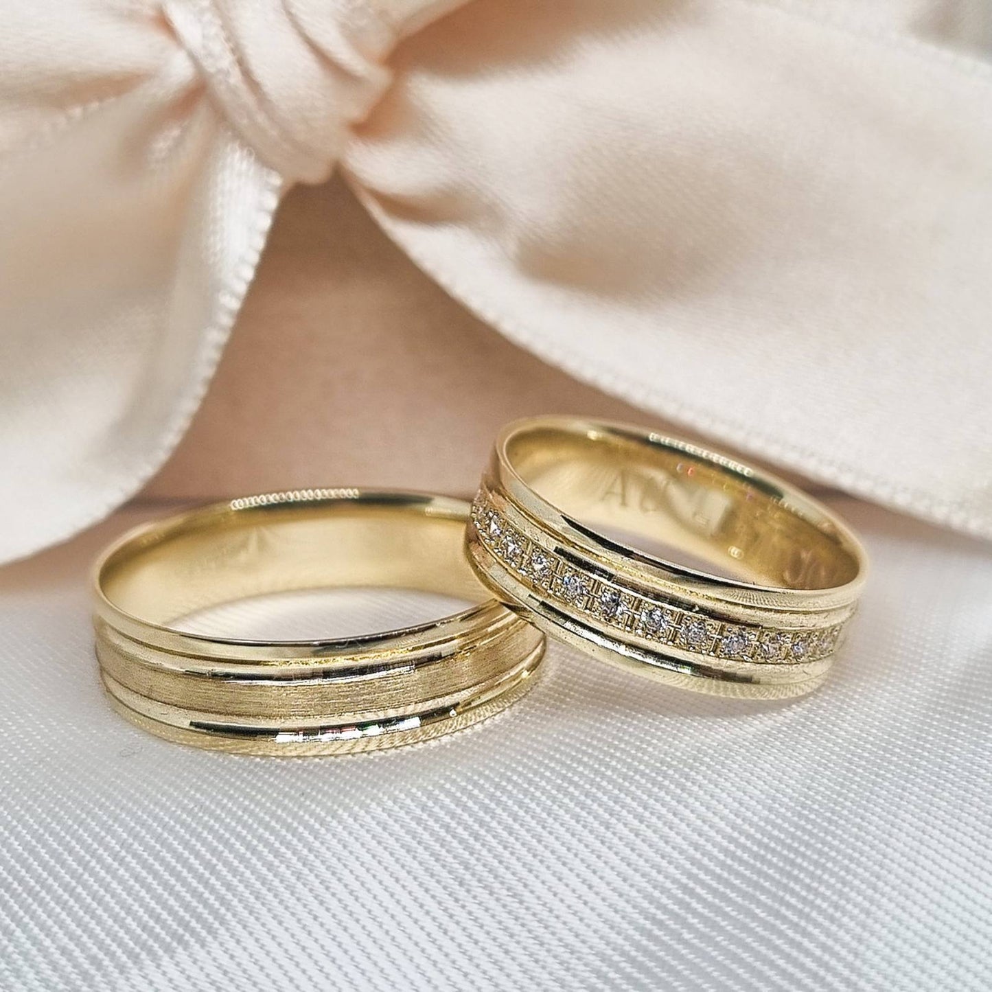 Zlatne burme od žutog zlata sa urezanim linijama, ženska burma dodatno ukrašena nizom sitnih cirkona, postavljene na belu tkaninu uz svetlu pozadinu sa mašnom.