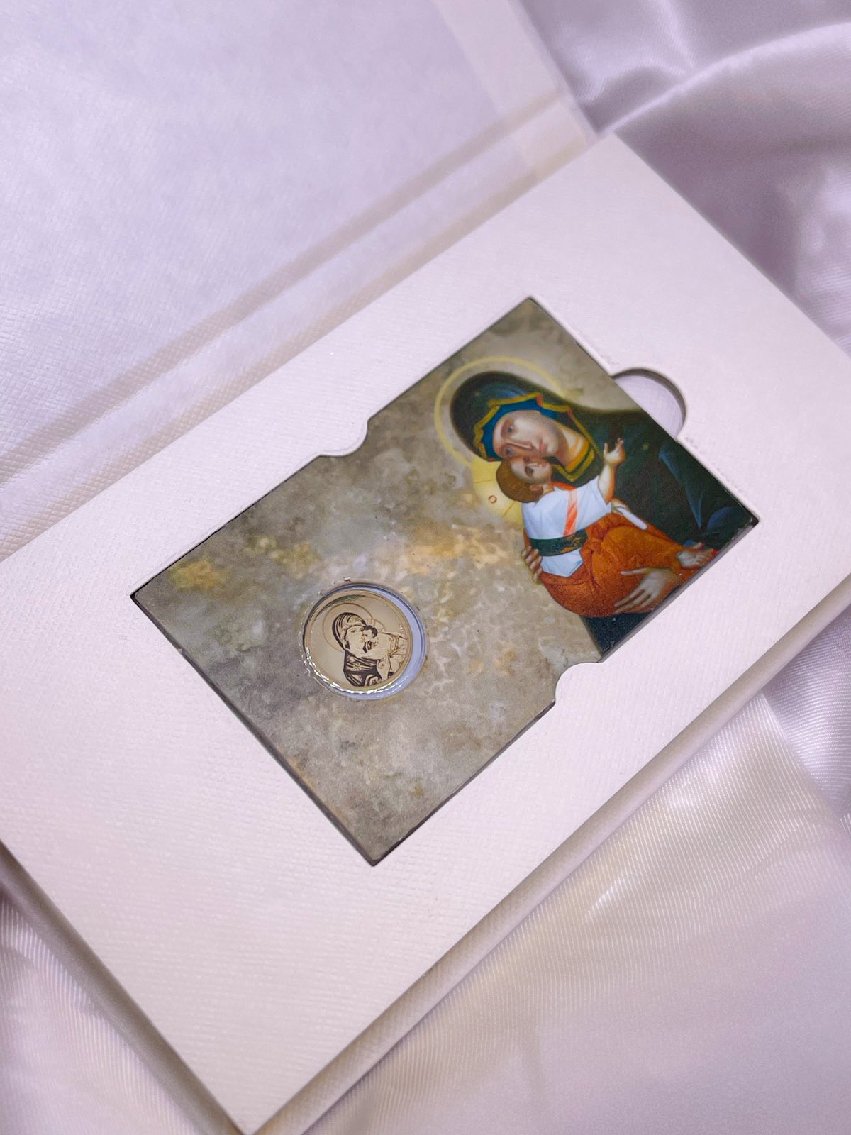 Zlatnik sa ugraviranim likom Bogorodice u blister pakovanju, uz prikaz ikone u boji na svetlucavoj podlozi, smešten u svetlu kutiju od teksturiranog papira.