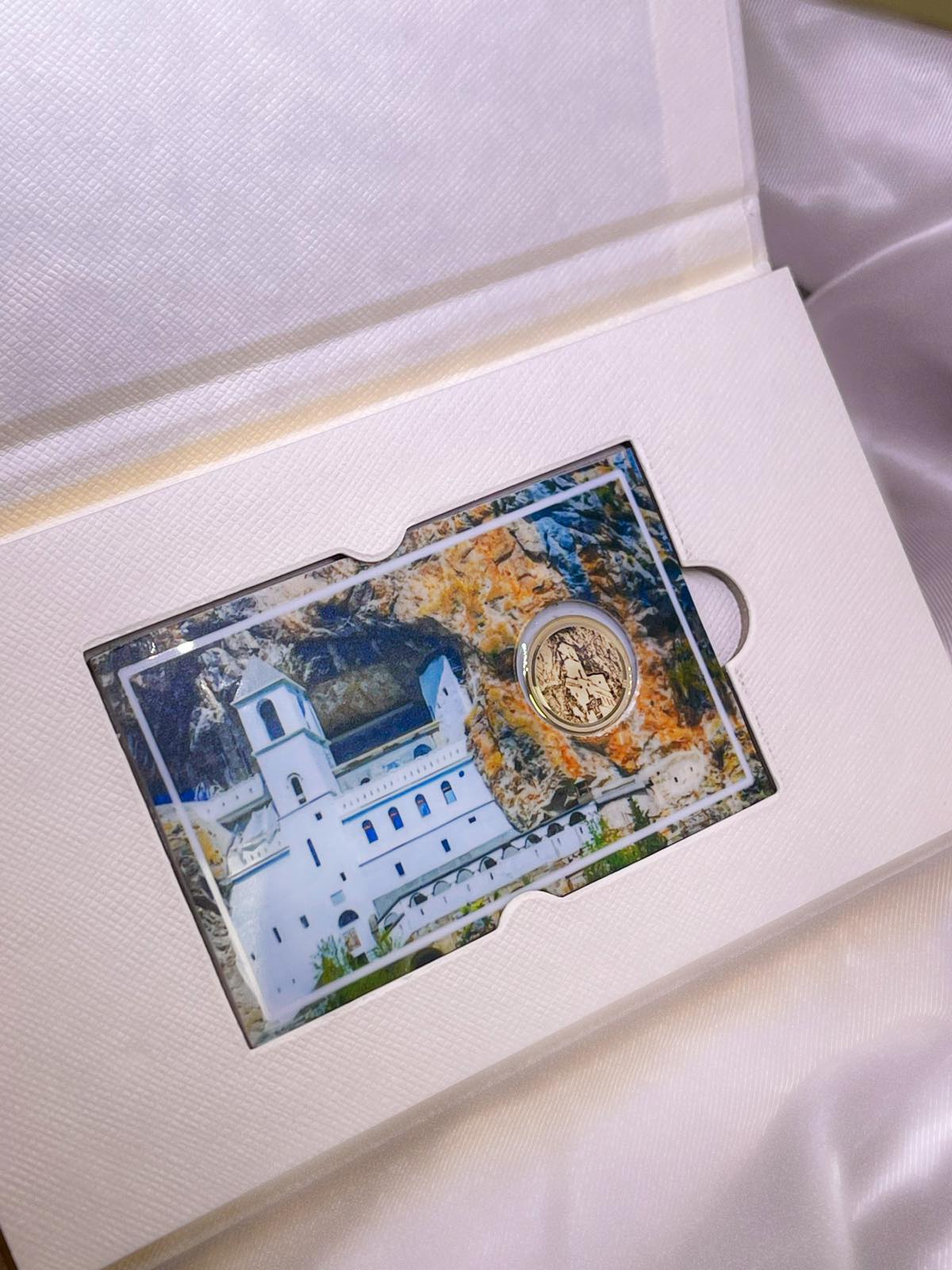 Zlatnik sa ugraviranim motivom Ostroga, smešten u blister sa pozadinom fotografije manastira uklesanog u stenu, u beloj kutiji sa teksturom.