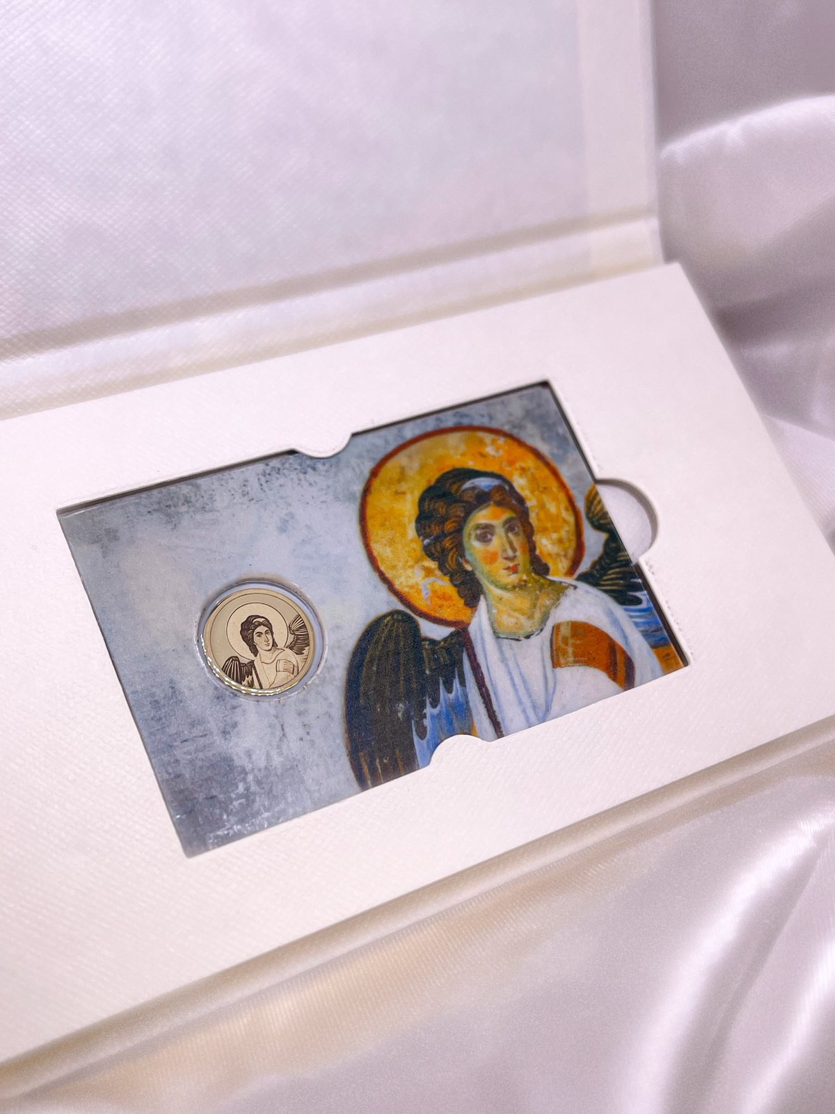 Zlatnik sa ugraviranom figurom Belog Anđela, postavljen u blister sa pozadinom freske iz manastira Mileševa, unutar svetle kutije sa svilenom podlogom.