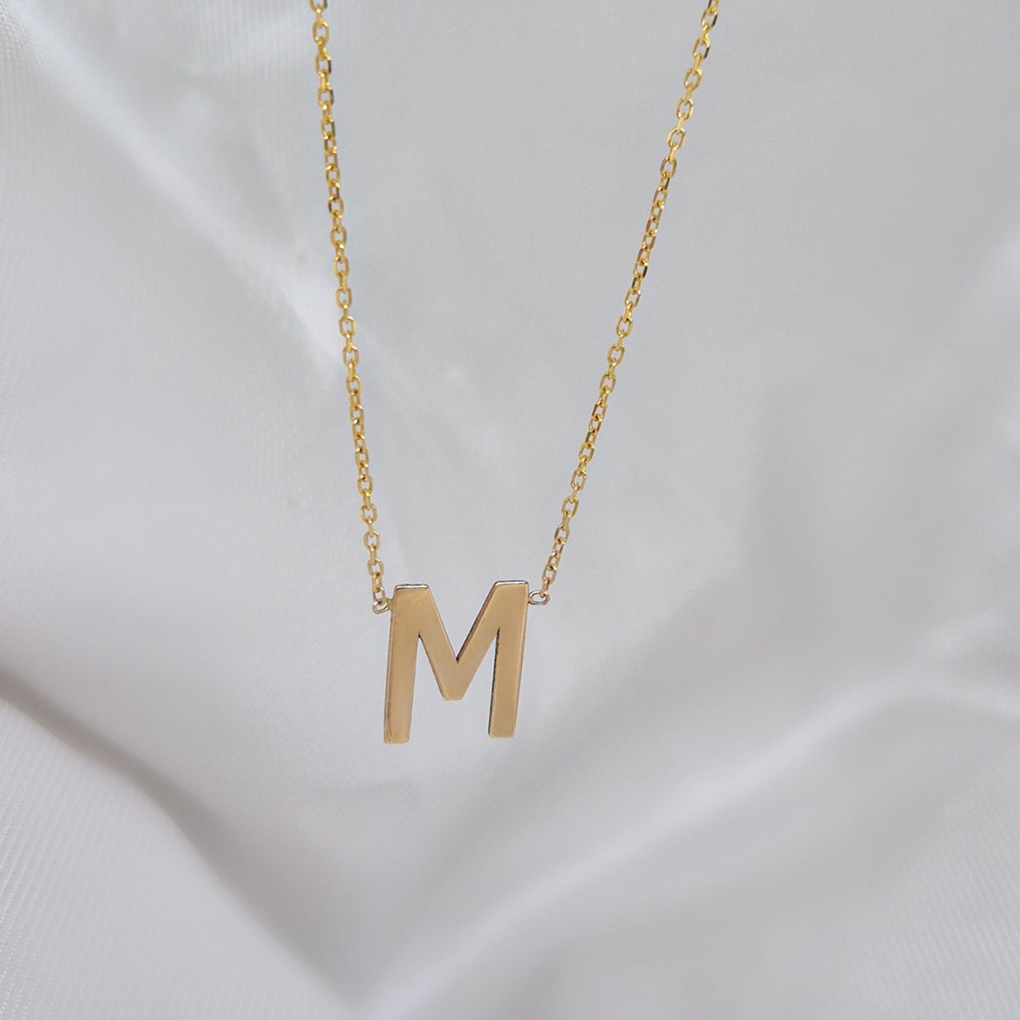 Zlatna ogrlica sa velikim slovom M na beloj tkanini