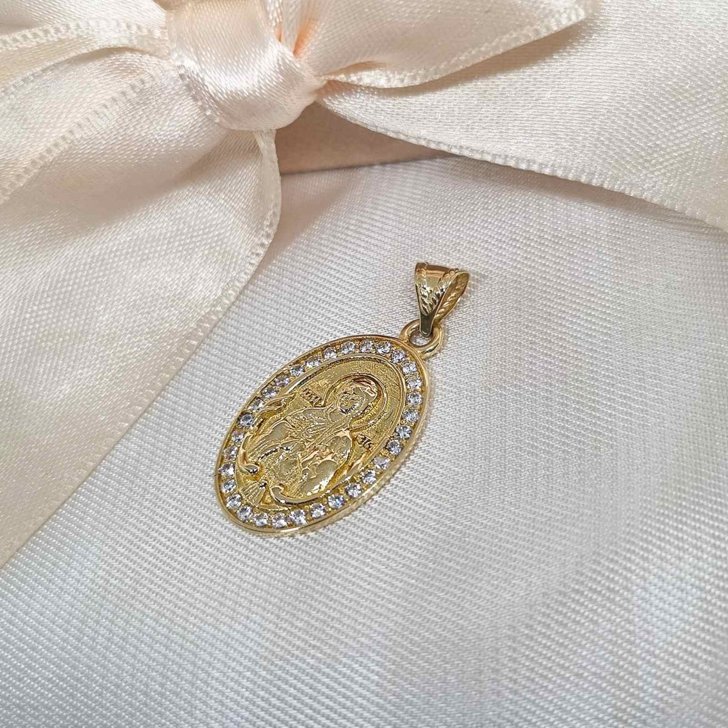Zlatni ovalni privezak sa Bogorodicom ugraviranom u sredini, okružen svetlucavim cirkonima, postavljen na belu tkaninu.
