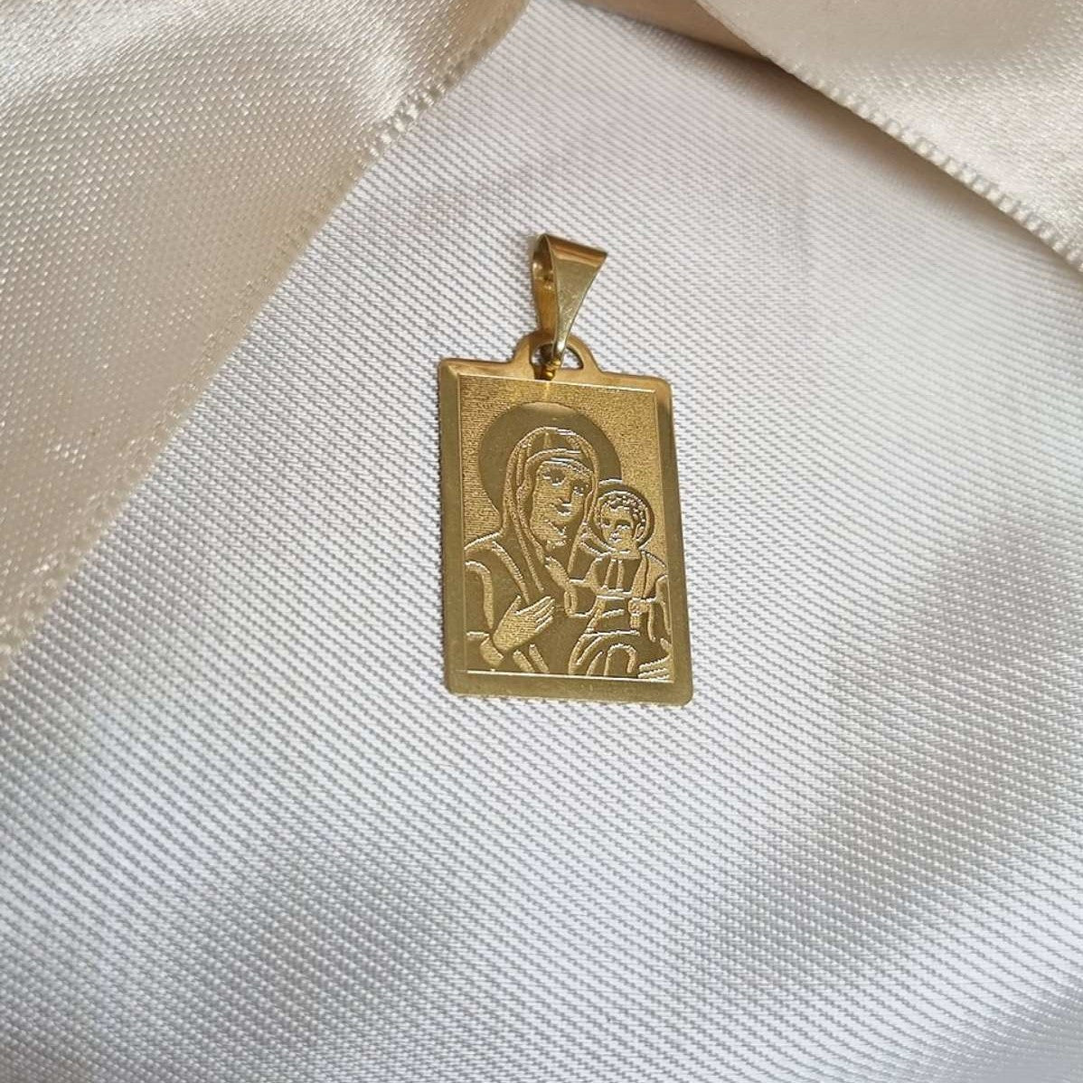 Zlatni pravougaoni privezak sa ugraviranim likom Bogorodice koja drži dete, postavljen na belu svilu.