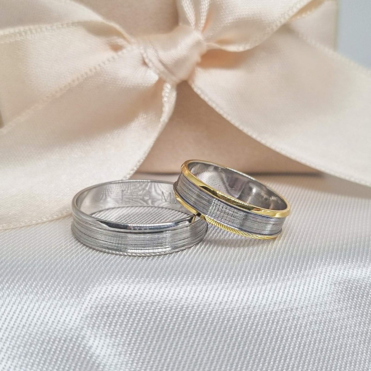 Par zlatnih burmi, jedna sa jednostavnim sjajnim dizajnom, a druga sa zlatnom ivicom i teksturiranim srednjim delom, postavljene na svilenkastoj pozadini u bež boji sa svetlom.