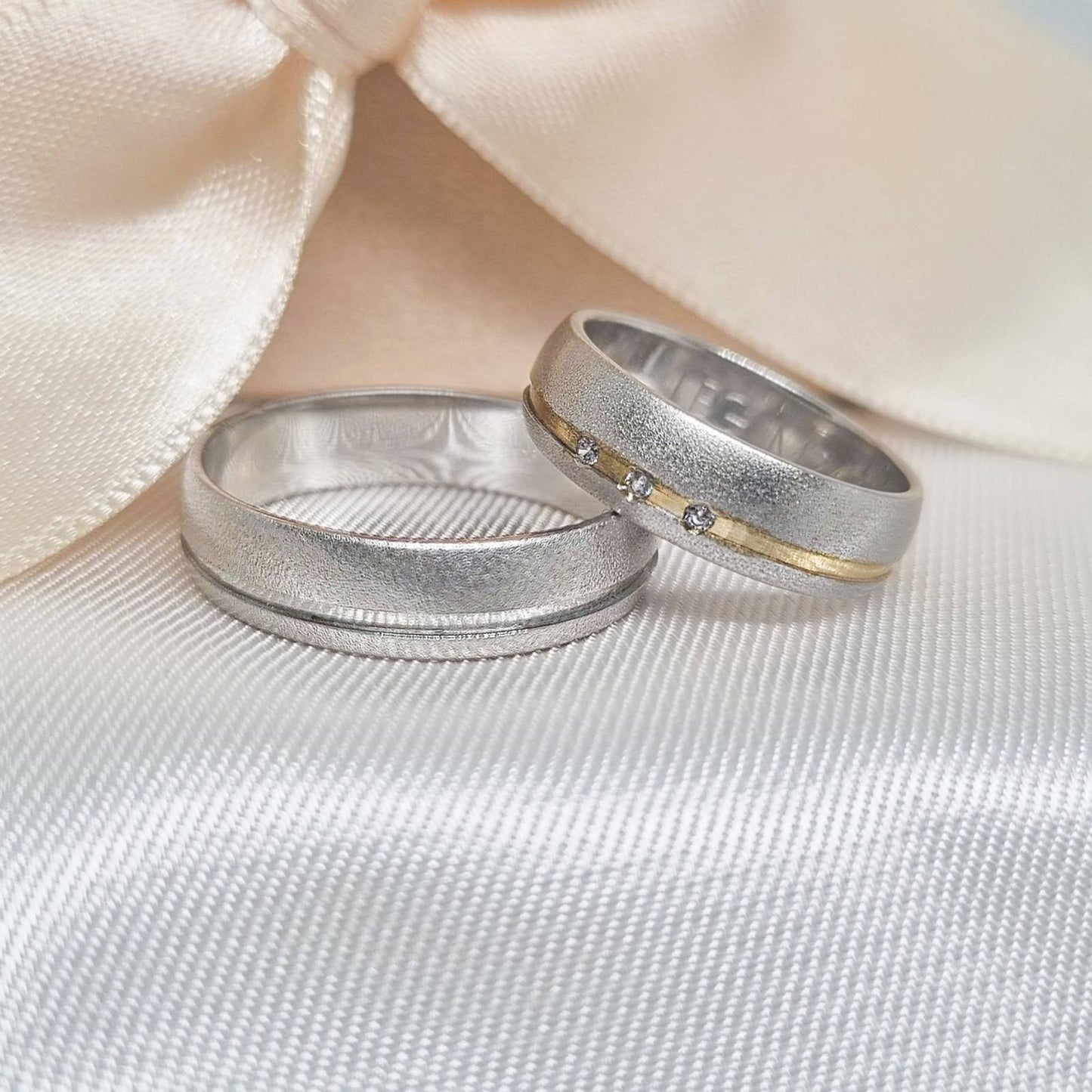 Par zlatnih burmi od belog zlata, muška sa diskretnim mat efektom, a ženska sa zlatnom linijom i tri cirkona, položene na svetlu tkaninu sa satenskom pozadinom.