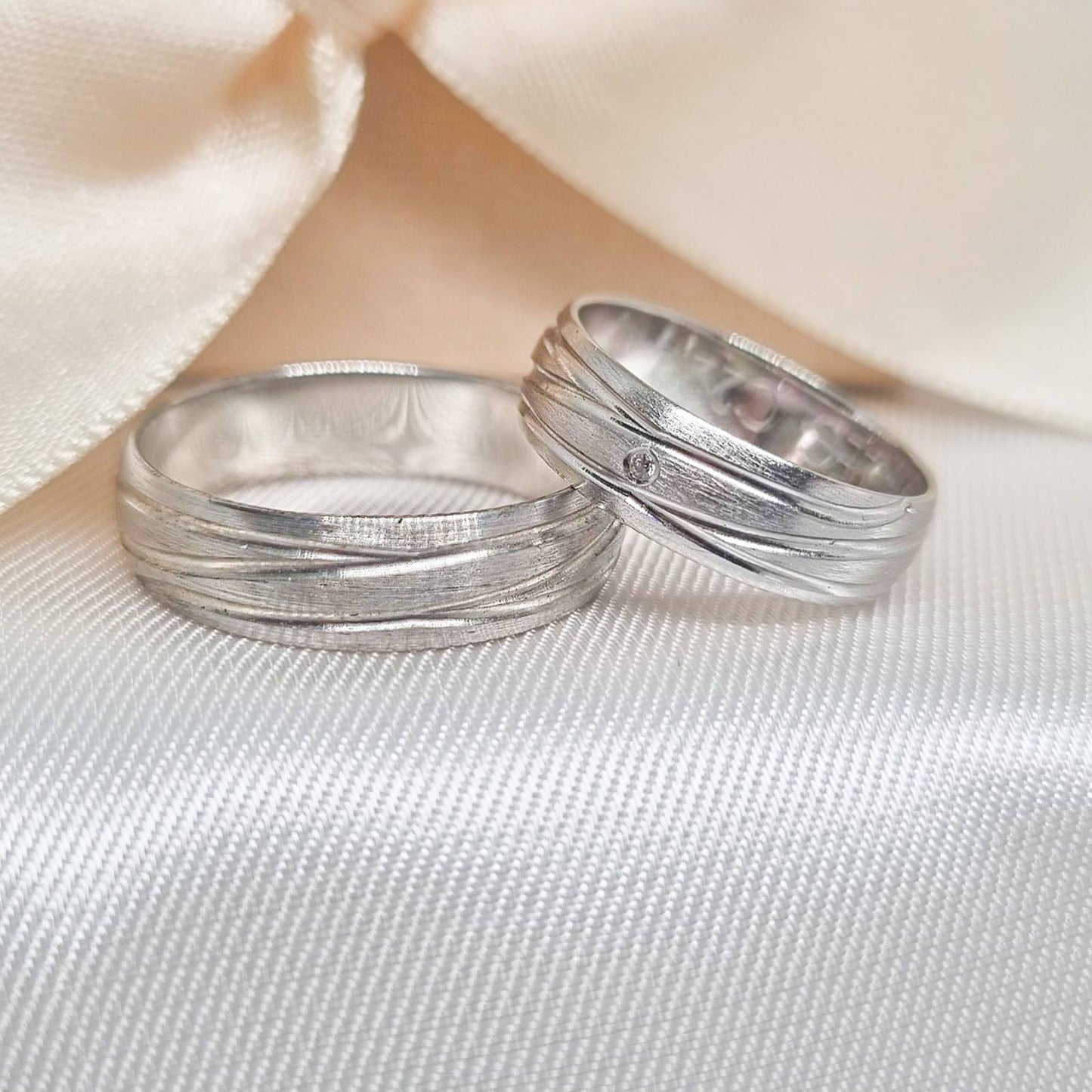 Zlatne burme od belog zlata sa talasastom teksturom, jedna ukrašena cirkonom, postavljene na svetloj tkanini sa bež pozadinom.