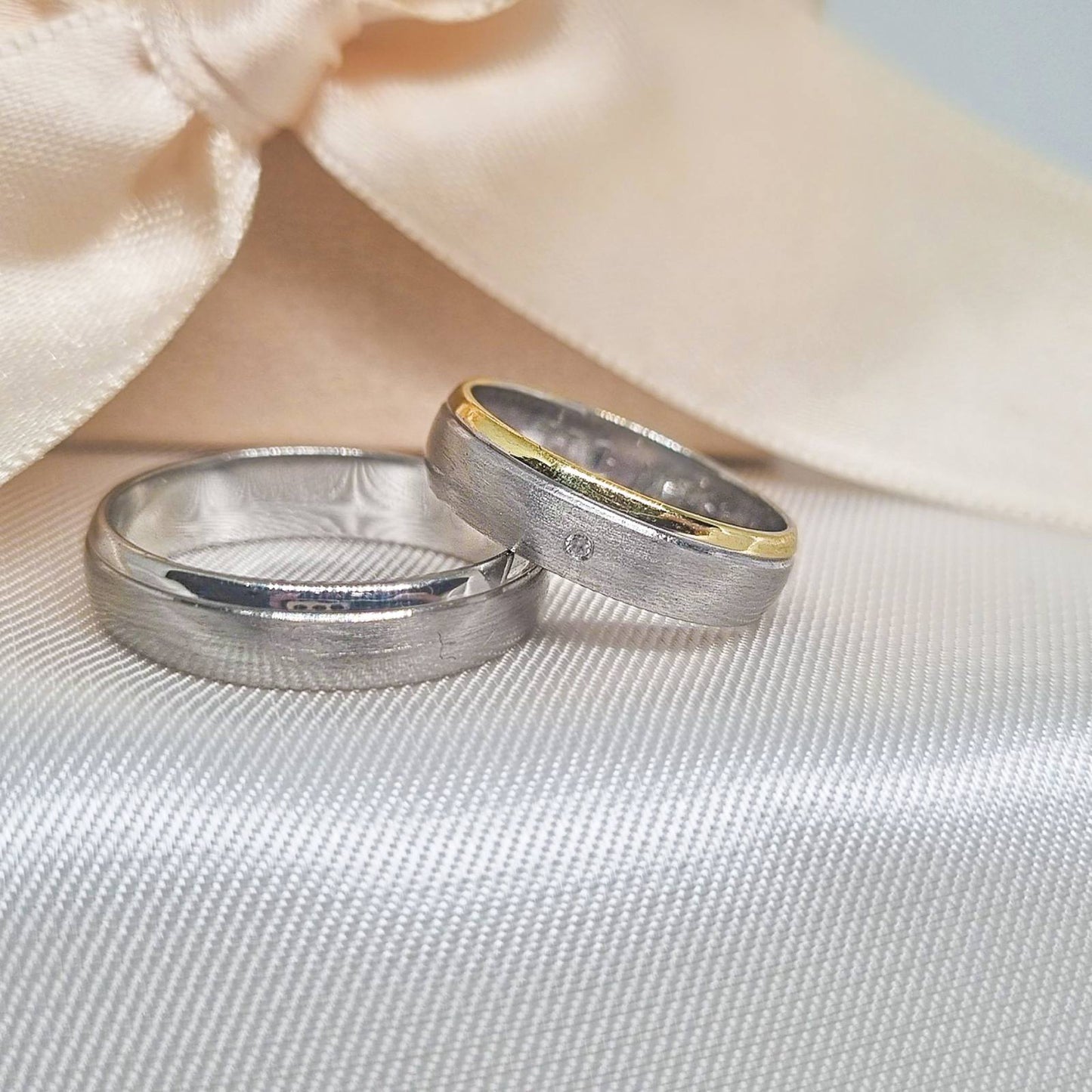 Zlatne burme sa matiranom površinom, jedna u potpunosti od belog zlata, a druga sa tankim žutim zlatnim prstenom na ivici, postavljene na mekoj bež tkanini sa svetlom pozadinom.