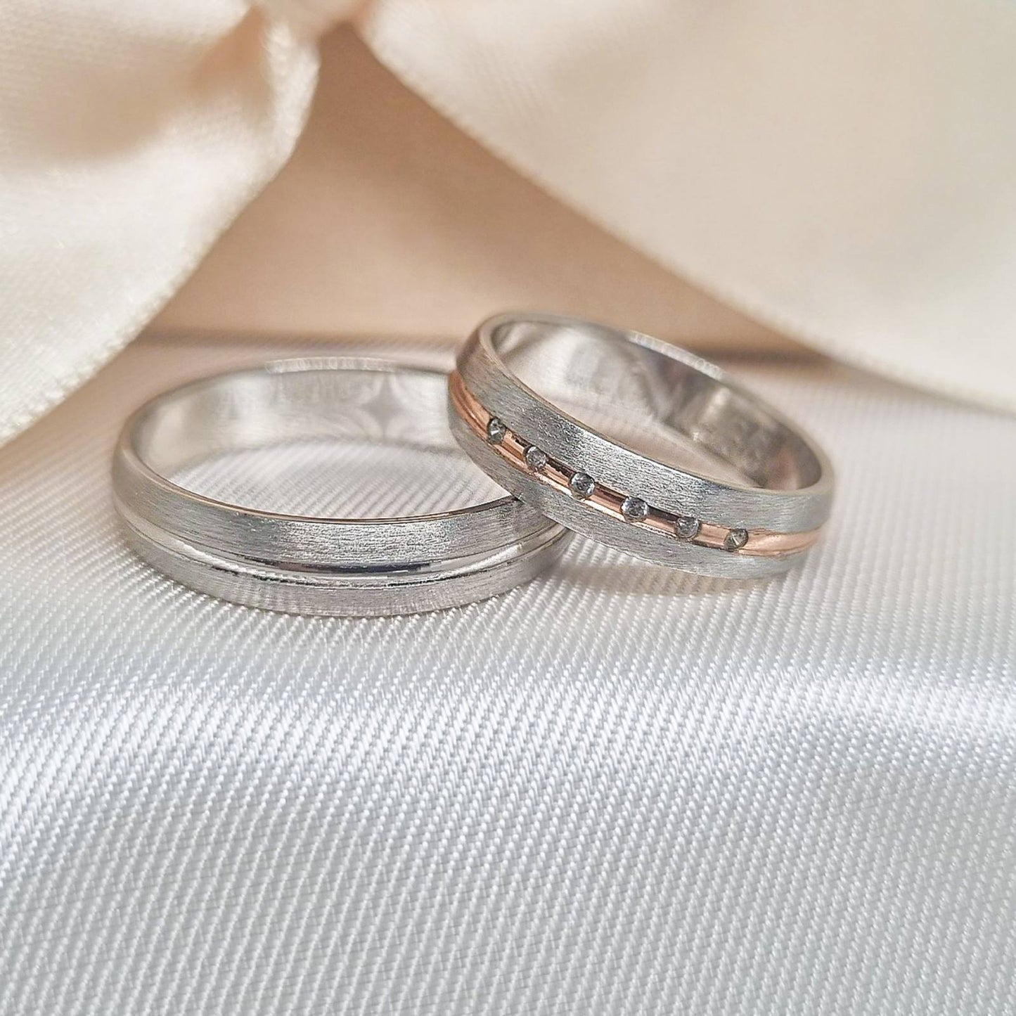 Burme od belog zlata sa satenskim finišem, jedna ukrašena roze zlatnom linijom i cirkonima, predstavljene na svetlom tekstilnom postavu ispred dekorativne mašne.