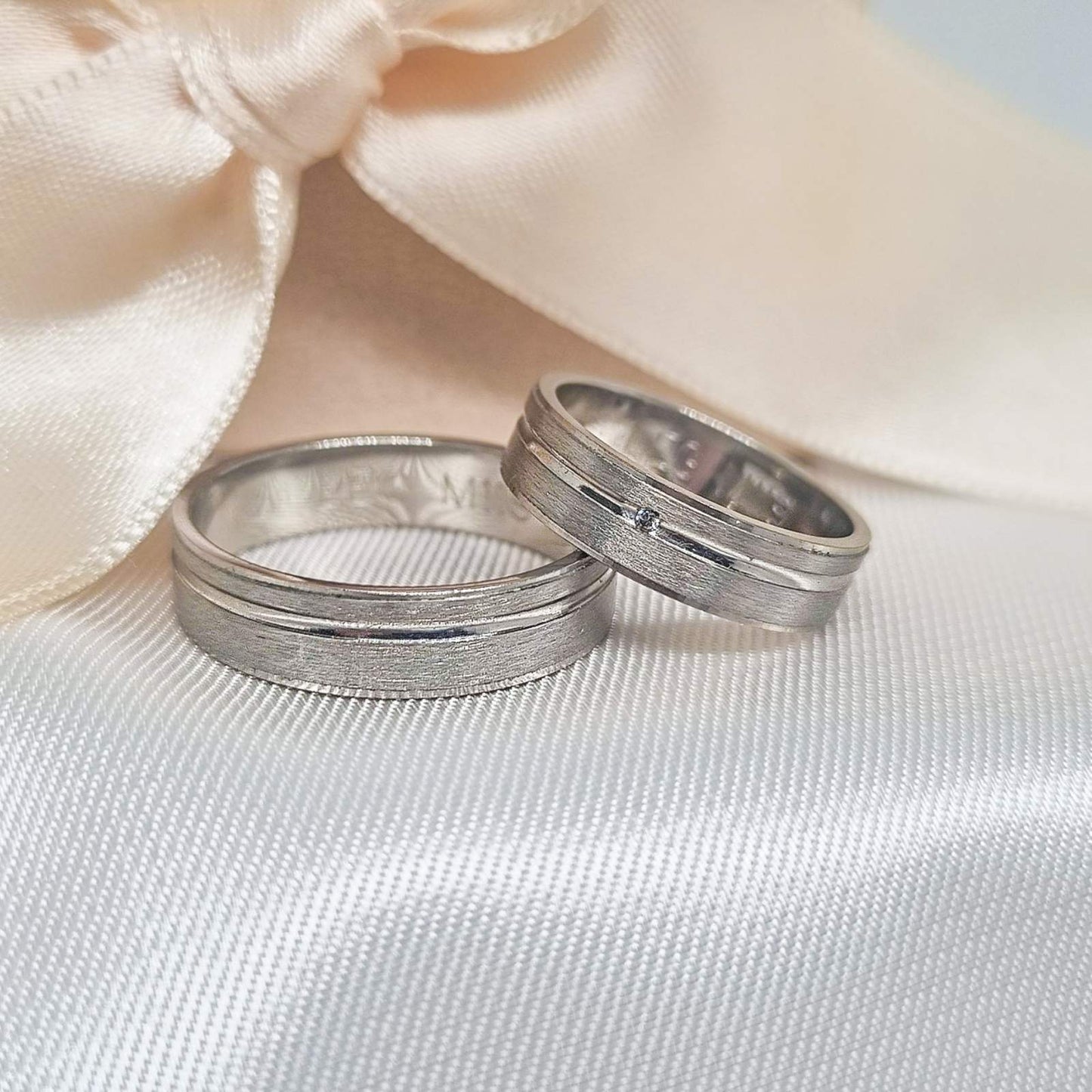 Zlatne burme sa matiranim i poliranim prugama, jedna sa malim dijamantom, postavljene na bež tkanini sa svetlom pozadinom.